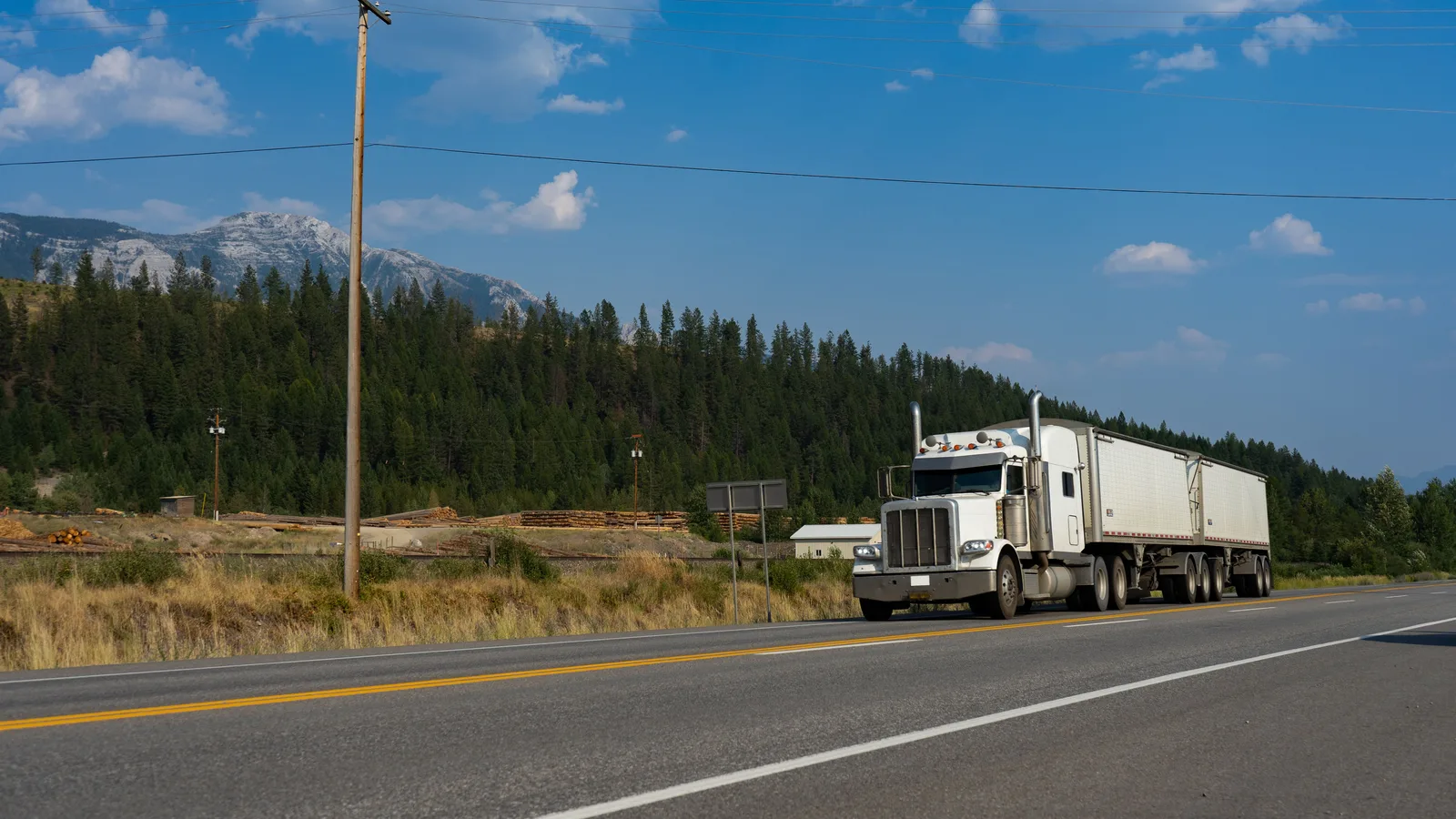 A white semi-truck pulling two trailers on a two-lane highway in the BC Interior, with forested hills and a mountain behind