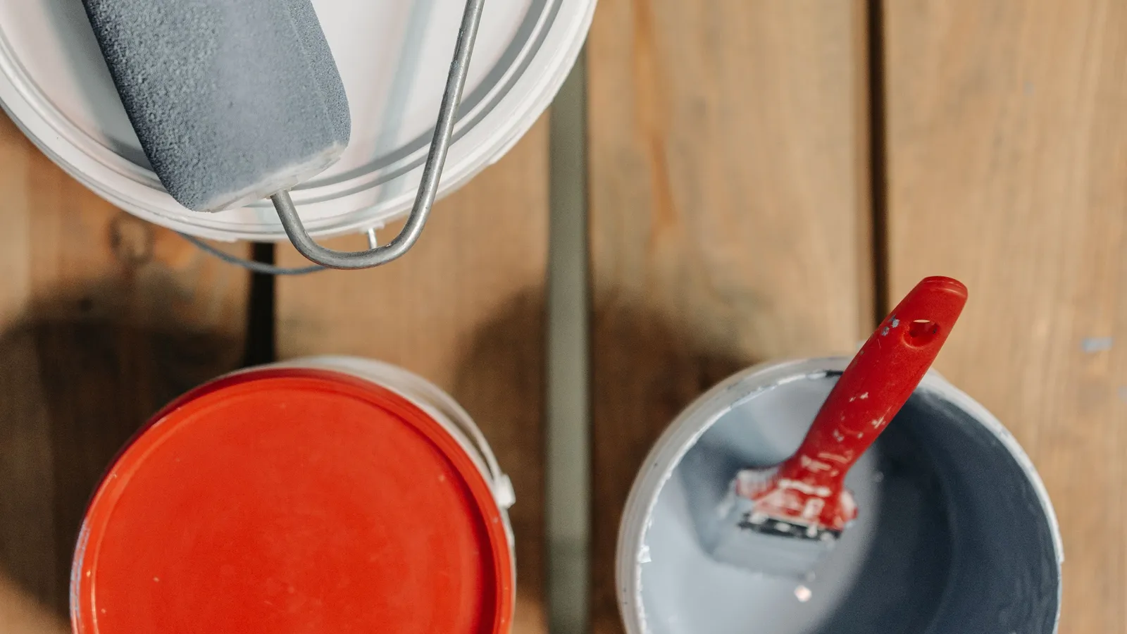 Top-down view of three paint cans on wooden boards, one with a paint roller resting on its lid and one open with a red-handled brush inside