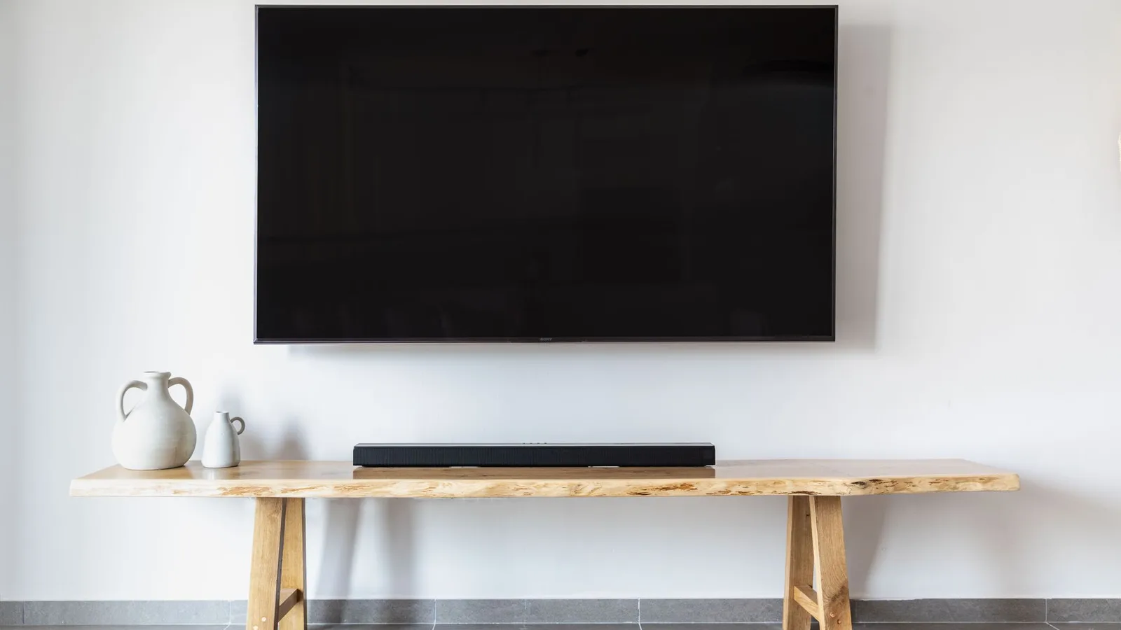 A black flat-screen TV mounted on a white wall above a long wooden console with a soundbar and two white vases